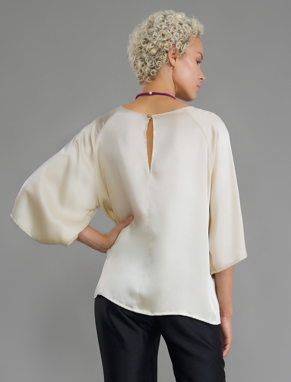 Back view. Lightweight 100% silk satin top in ivory colour, features a wide neckline with a button fastening at the back, a gently curved hemline and wide sleeves 3/4 length sleeves.