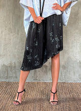 Load image into Gallery viewer, This asymmetric Skirt-Pant is handcrafted from silk satin and blur the line between shorts and skirt. A deep, gathered waistband flows into a fluid silhouette, finished with a diagonal hemline that brings an unconventional edge to refined dressing.This  hand-drawn batik motif is inspired from the late 19th-century ‘Wiener Secession’ art movement, infusing the design with a refined sense of linear rigor softened by small foliage details. here in black background and steel grey motif.
