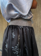 Load image into Gallery viewer, Elasticated waist detail:this asymmetric Skirt-Pant is handcrafted from silk satin and blur the line between shorts and skirt. A deep, gathered waistband flows into a fluid silhouette, finished with a diagonal hemline that brings an unconventional edge to refined dressing.This  hand-drawn batik motif is inspired from the late 19th-century ‘Wiener Secession’ art movement, infusing the design with a refined sense of linear rigor softened by small foliage details. here in black background and steel grey motif.
