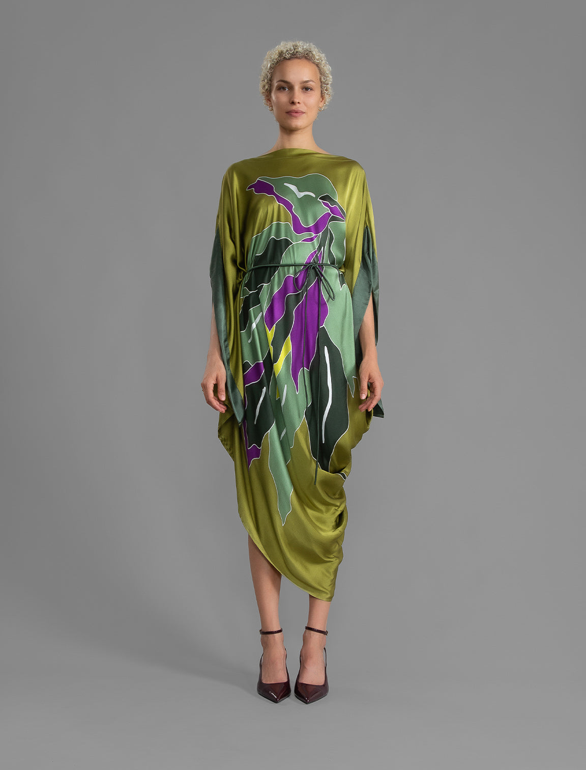 Front view with belt. Elegant silk kaftan dress with oversized pink floral batik motif on deep vino color silk, featuring flowing kimono sleeves, asymmetrical hemline, and relaxed contemporary silhouette.
