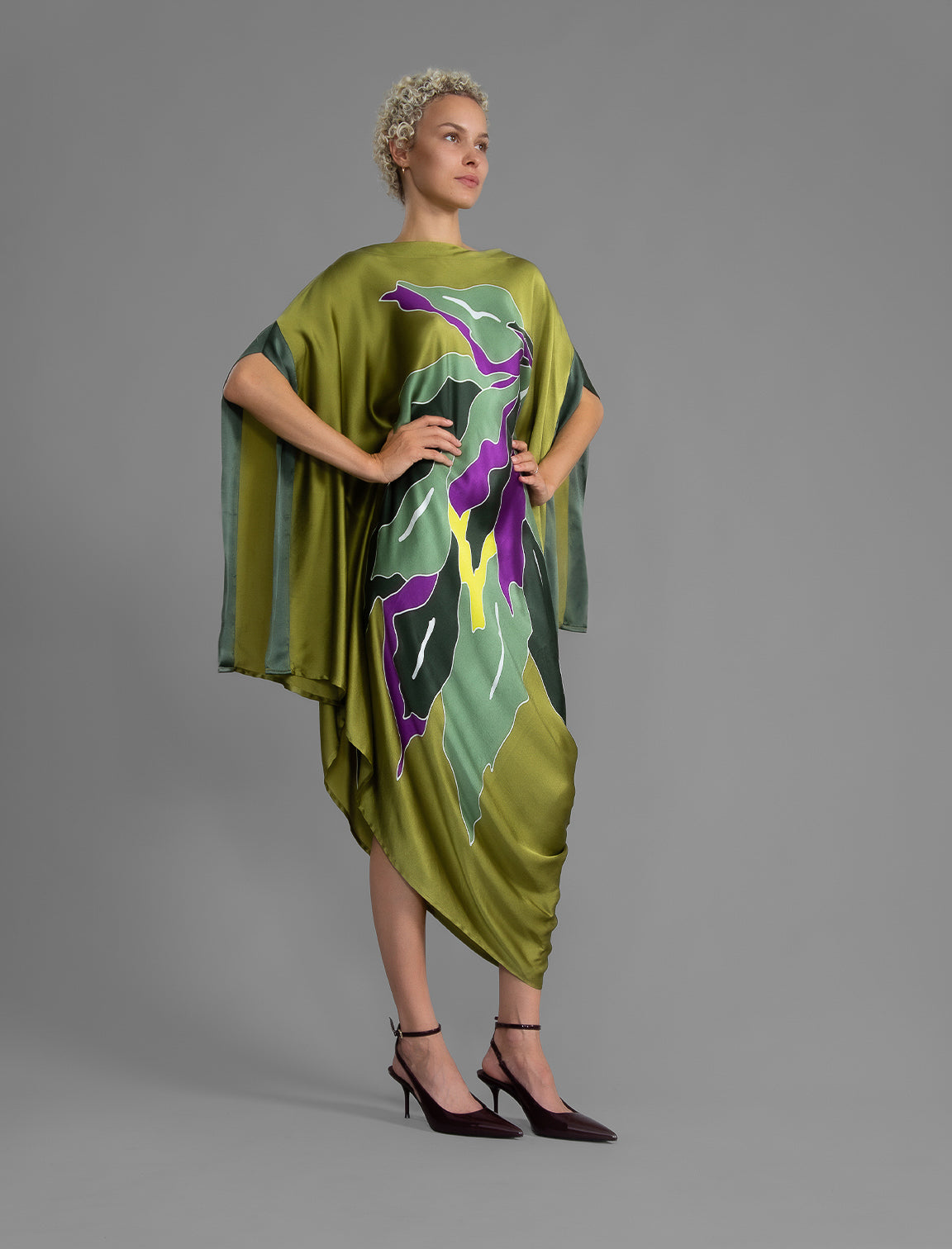 Side view. Elegant silk kaftan dress with oversized tropical leaf motif in moss green color silk, featuring flowing kimono sleeves, asymmetrical hemline, and relaxed contemporary silhouette.