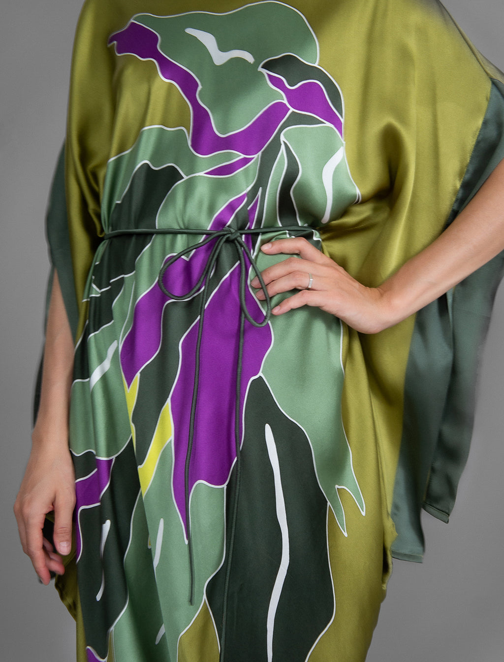 Detail view. Elegant silk kaftan dress with oversized tropical leaf motif in moss green color silk, featuring flowing kimono sleeves, asymmetrical hemline, and relaxed contemporary silhouette.
