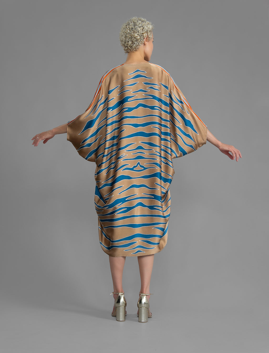 Back view. Relaxed flowing silk kaftan with graphic pale blue zebra motif on caramel colour, with orange detail, featuring 3/4 length sleeves and a voluminous silhouette.