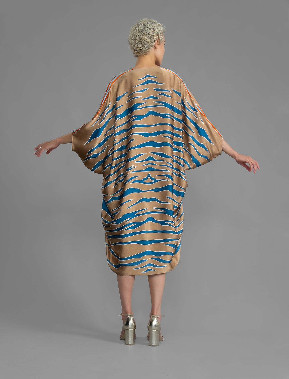Back view. Relaxed flowing silk kaftan with graphic pale blue zebra motif on caramel colour, with orange detail, featuring 3/4 length sleeves and a voluminous silhouette.
