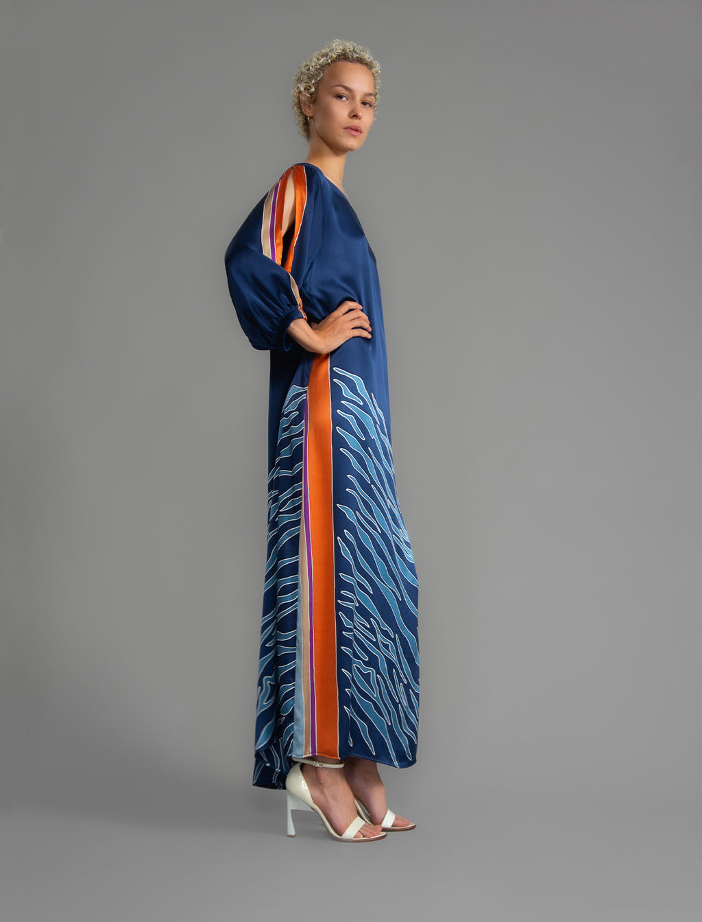 Side view. Full length 100% silk tunic dress with blue zebra motif and orange details on navy blue colour, featuring a open, wide neckline and one side split on the skirt, gathered 3/4 length sleeves with a slash opening and optional ribbon belt.