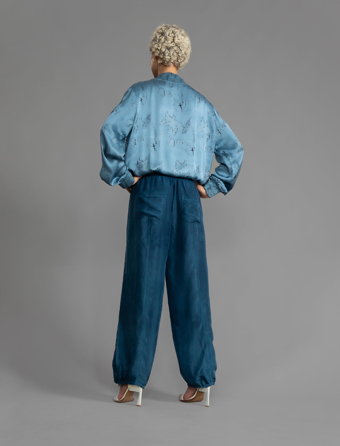 Back view. Super lightweight 100% habutai silk pant in soft mid blue, with wide leg, drawstring gather at the ankle, a deep jersey waistband with a drawstring and patch pockets on the back.
