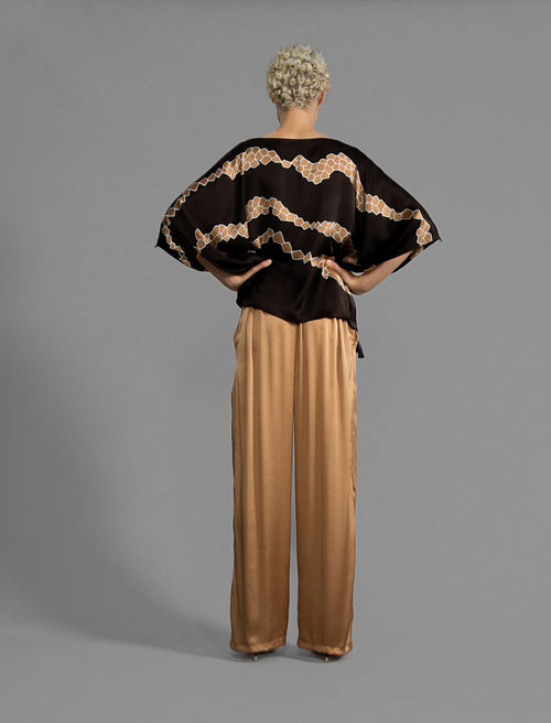 Back view. Relaxed wide-leg silk trousers in warm tobacco, featuring drawstring waistband, elasticated at the back. Soft gathered drape, fluid silhouette, and subtle lustrous finish.