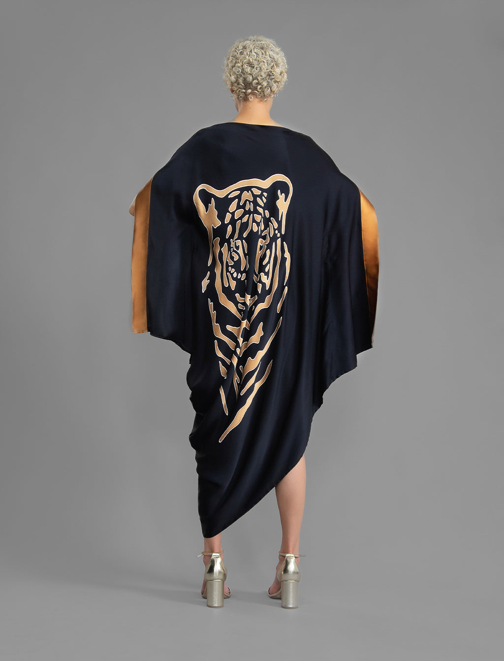 Back view. Elegant silk kaftan dress with tiger head batik motif on black color silk, featuring flowing kimono sleeves, asymmetrical hemline, and relaxed contemporary silhouette.