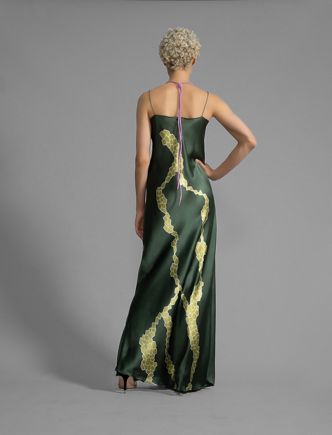 Back view. Elegant long body contouring silk dress in forest green colour with contrasting green graphic Macrosnake batik motif, featuring spaghetti straps, V-neckline and a fluid contemporary silhouette.