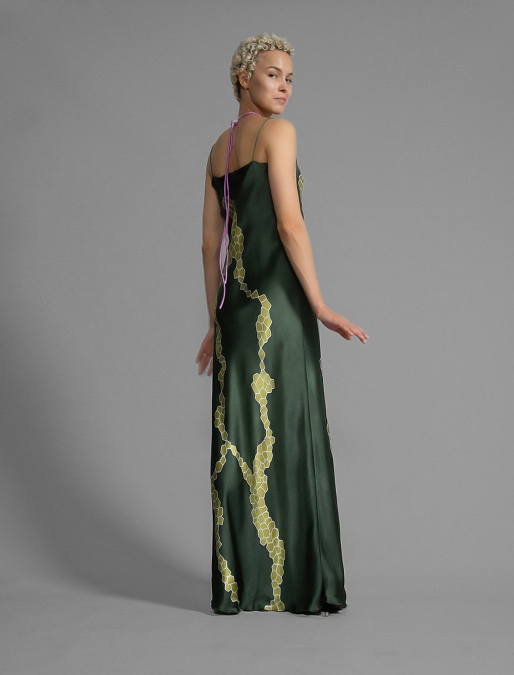Side view. Elegant long body contouring silk dress in forest green colour with contrasting green graphic Macrosnake batik motif, featuring spaghetti straps, V-neckline and a fluid contemporary silhouette.