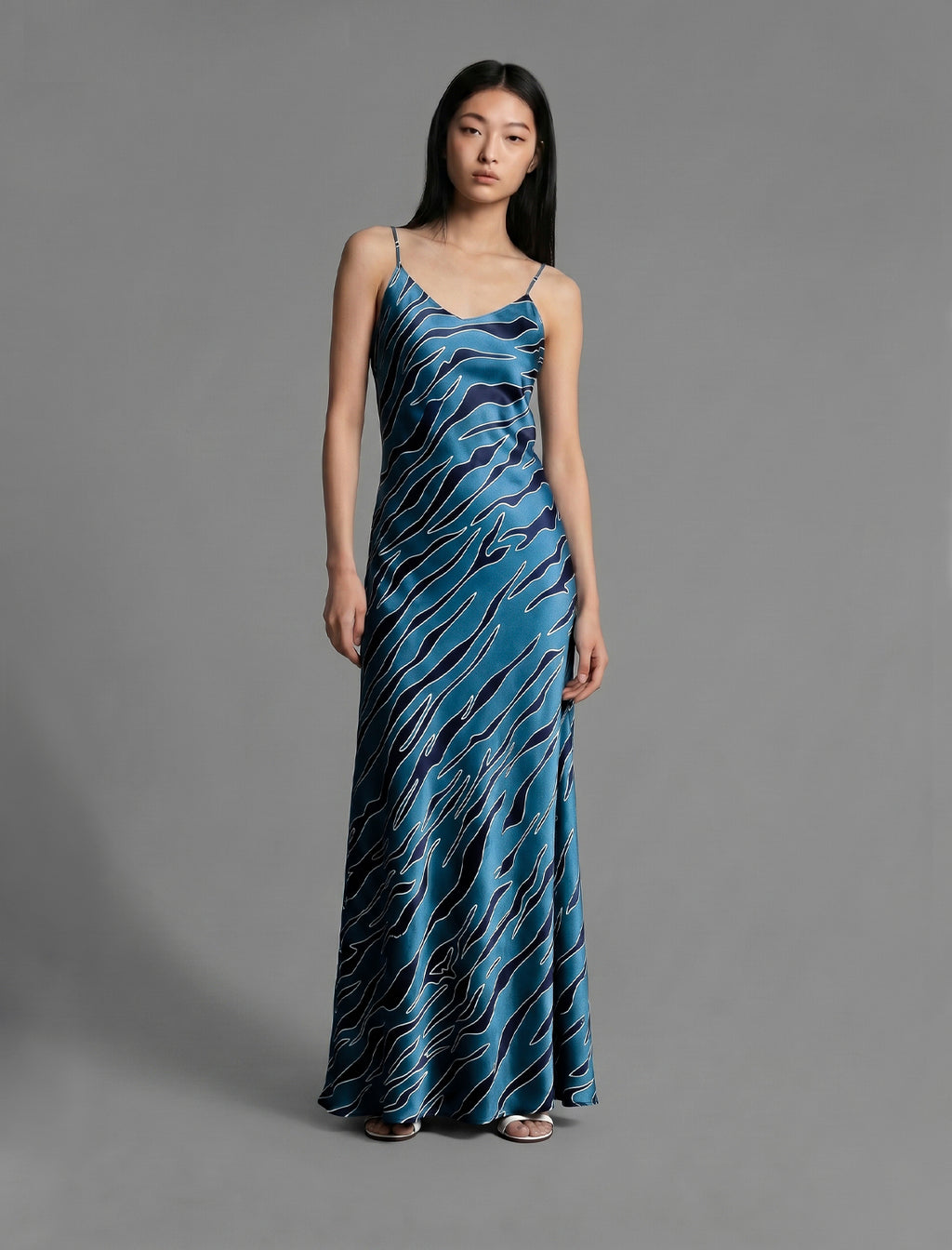Front view. Elegant long body contouring silk dress in blue colour with contrasting deep blue graphic Zebra batik motif, featuring spaghetti straps, V-neckline and a fluid contemporary silhouette.