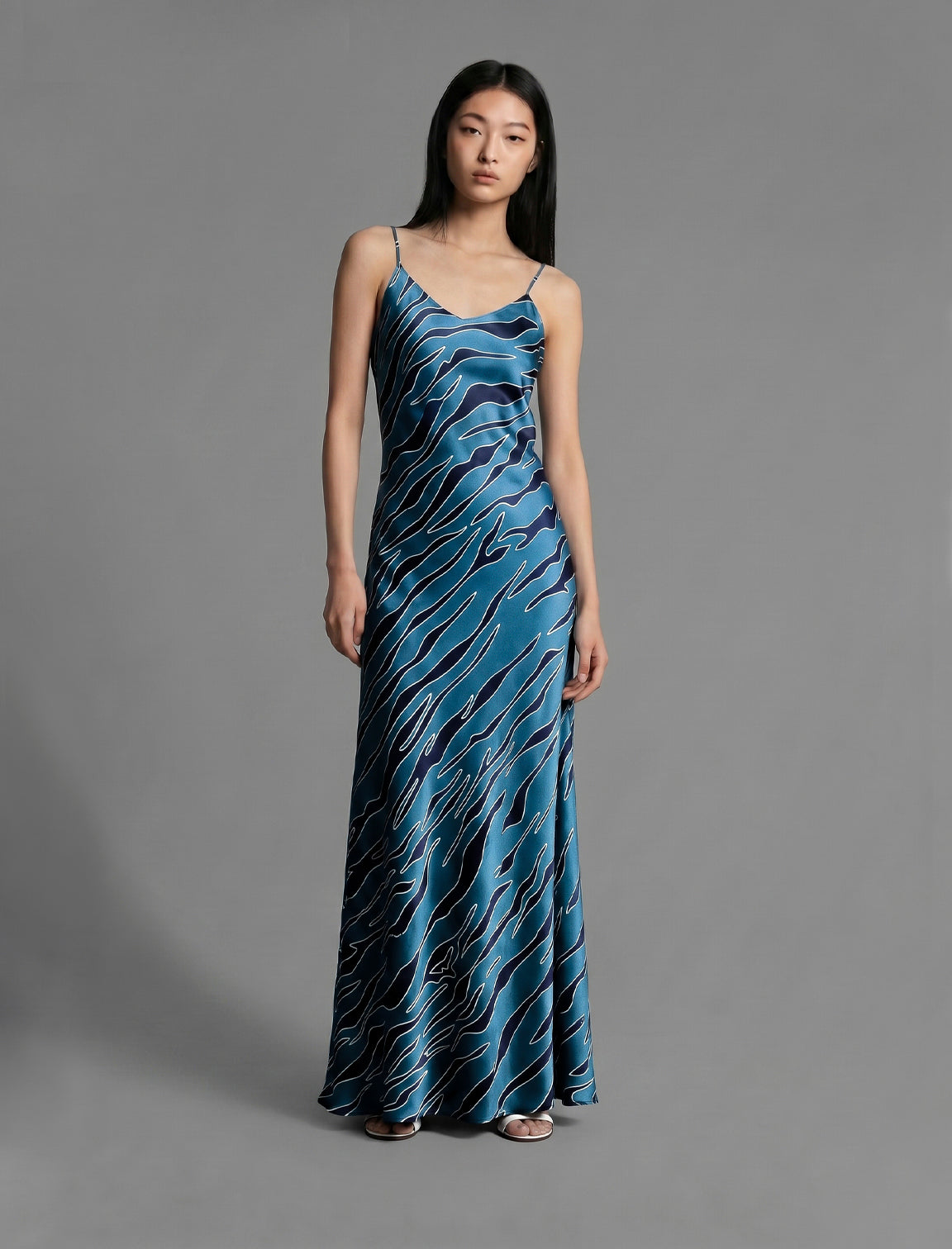 Front view. Elegant long body contouring silk dress in blue colour with contrasting deep blue graphic Zebra batik motif, featuring spaghetti straps, V-neckline and a fluid contemporary silhouette.