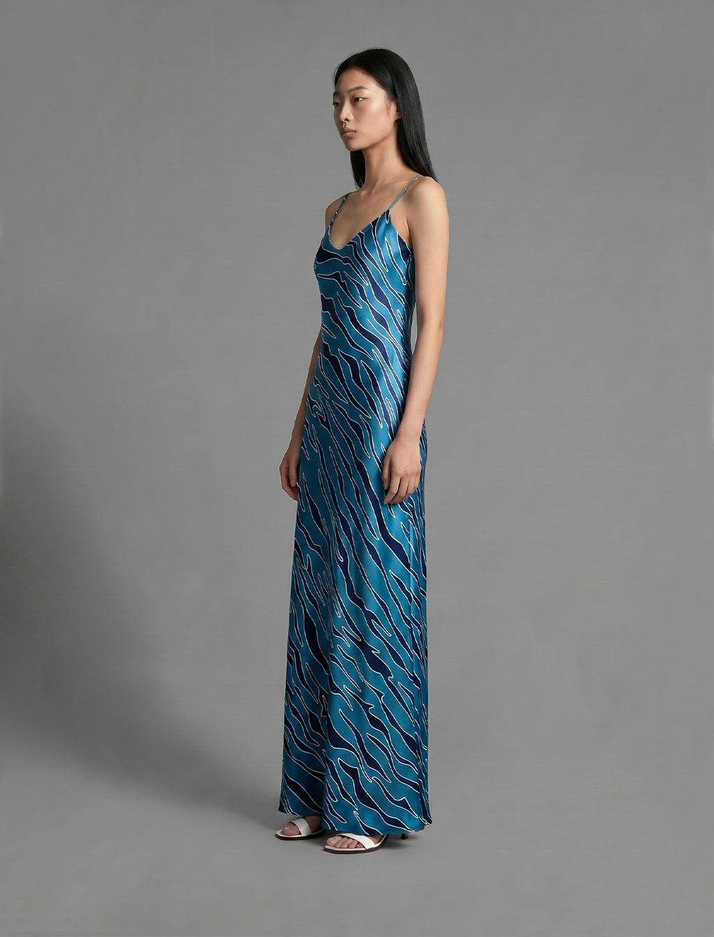 Side view. Elegant long body contouring silk dress in blue colour with contrasting deep blue graphic Zebra batik motif, featuring spaghetti straps, V-neckline and a fluid contemporary silhouette.