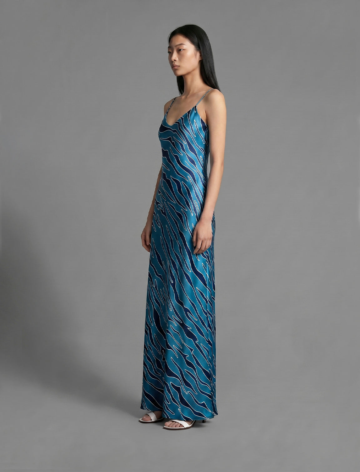 Side view. Elegant long body contouring silk dress in blue colour with contrasting deep blue graphic Zebra batik motif, featuring spaghetti straps, V-neckline and a fluid contemporary silhouette.
