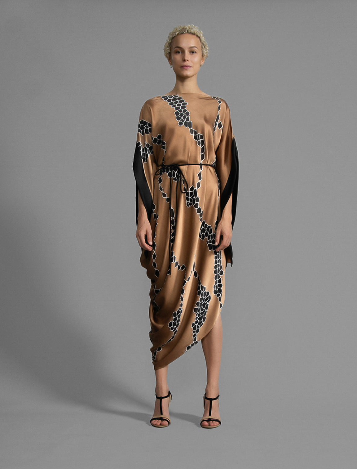 Front view. Elegant silk draped dress with graphic Macrosnake batik motif in black on warm tobacco colour, featuring fluid kimono sleeves, asymmetrical hemline, and relaxed contemporary silhouette with waist tie detail.