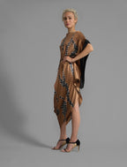Left side view. Elegant silk draped dress with graphic Macrosnake batik motif in black on warm tobacco colour, featuring fluid kimono sleeves, asymmetrical hemline, and relaxed contemporary silhouette with waist tie detail.