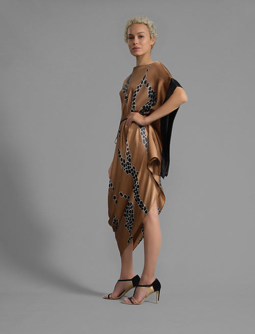 Left side view. Elegant silk draped dress with graphic Macrosnake batik motif in black on warm tobacco colour, featuring fluid kimono sleeves, asymmetrical hemline, and relaxed contemporary silhouette with waist tie detail.