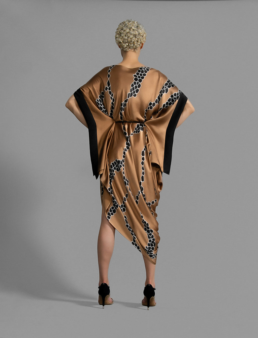 Back view. Elegant silk draped dress with graphic Macrosnake batik motif in black on warm tobacco colour, featuring fluid kimono sleeves, asymmetrical hemline, and relaxed contemporary silhouette with waist tie detail.