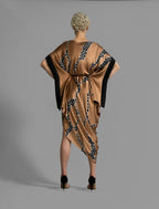 Back view. Elegant silk draped dress with graphic Macrosnake batik motif in black on warm tobacco colour, featuring fluid kimono sleeves, asymmetrical hemline, and relaxed contemporary silhouette with waist tie detail.