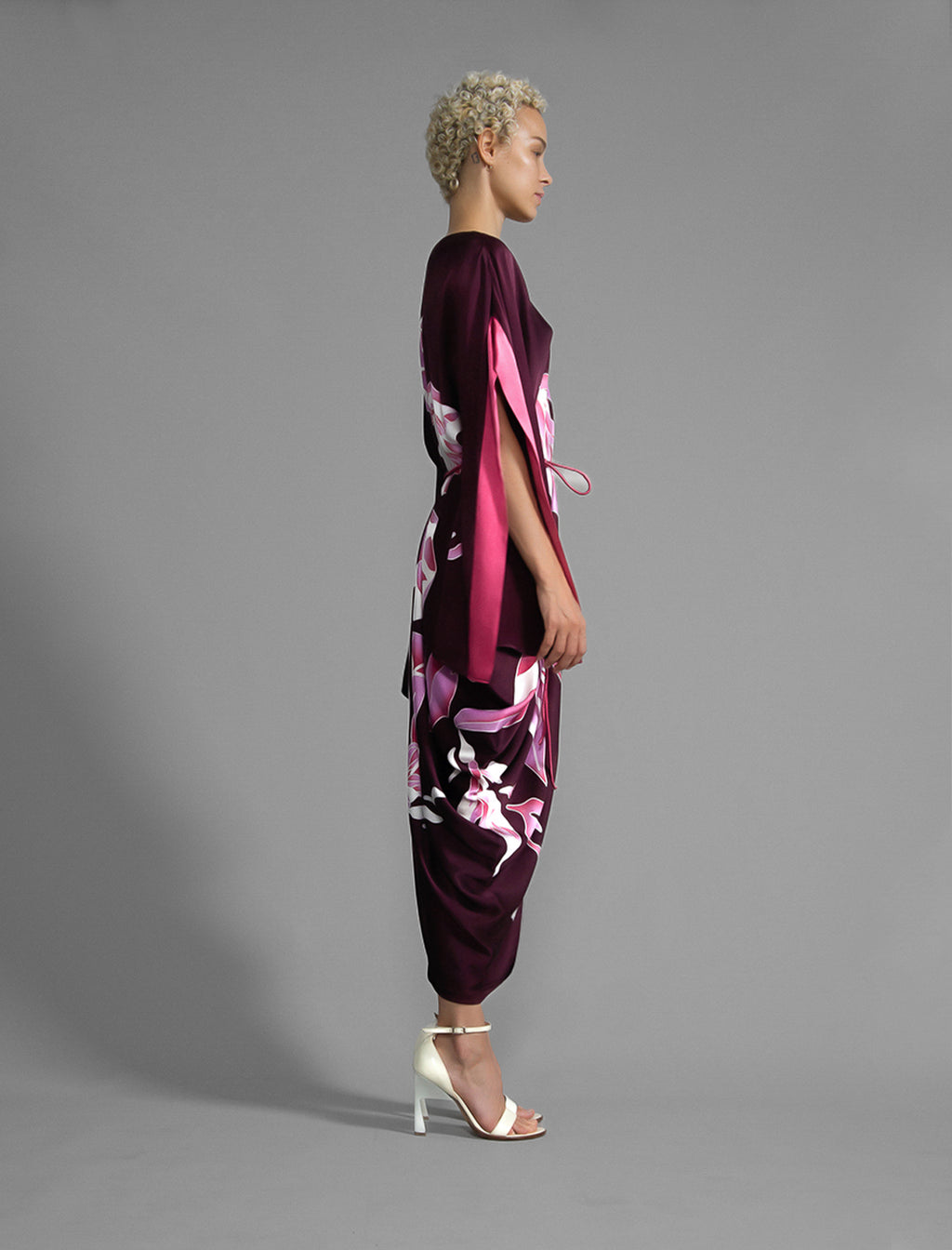 Side view. Elegant silk kaftan dress with oversized pink floral batik motif on deep vino colour silk, featuring flowing kimono sleeves, asymmetrical hemline, and relaxed contemporary silhouette.