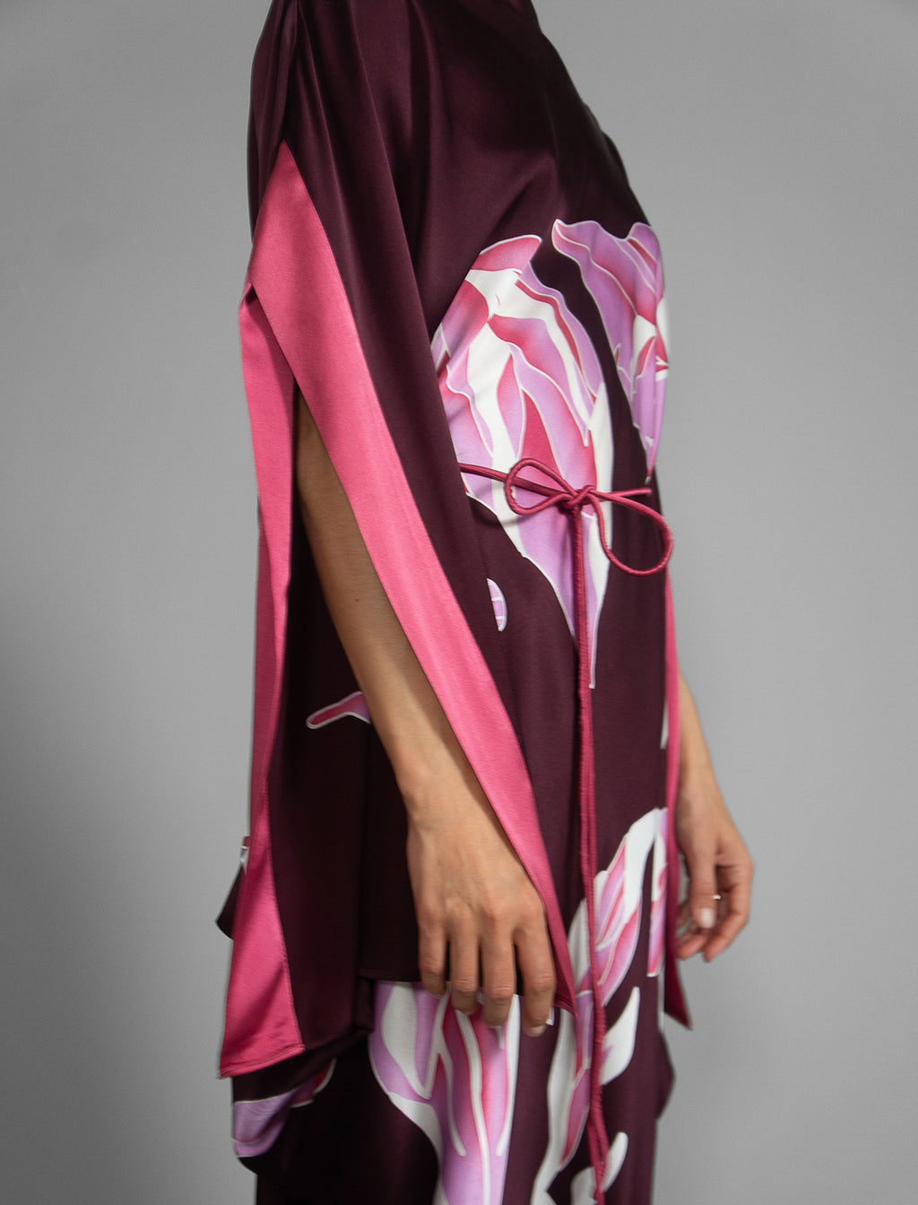 Detail view. Elegant silk kaftan dress with oversized pink floral batik motif on deep vino colour silk, featuring flowing kimono sleeves, asymmetrical hemline, and relaxed contemporary silhouette.