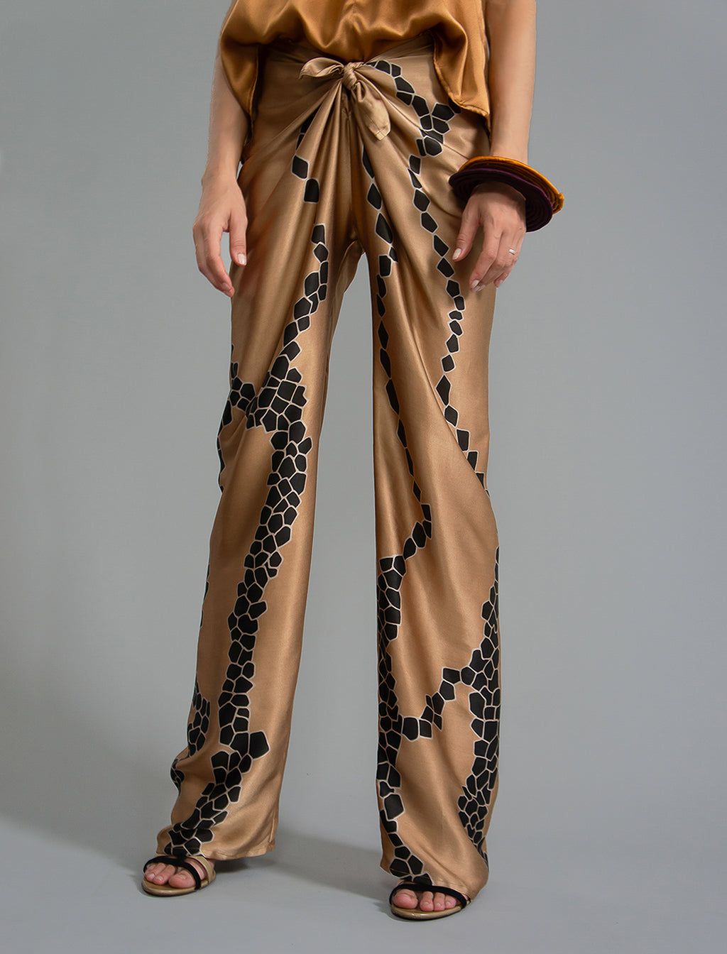 Close front view. Long silk pants in tobacco colour with contrasting blue graphic Macrosnake batik motif, featuring a knot tie at the front and a slick relaxed silhouette. 