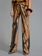 Front detail view. Long silk pants in tobacco colour with contrasting blue graphic Macrosnake batik motif, featuring a knot tie at the front and a slick relaxed silhouette. 