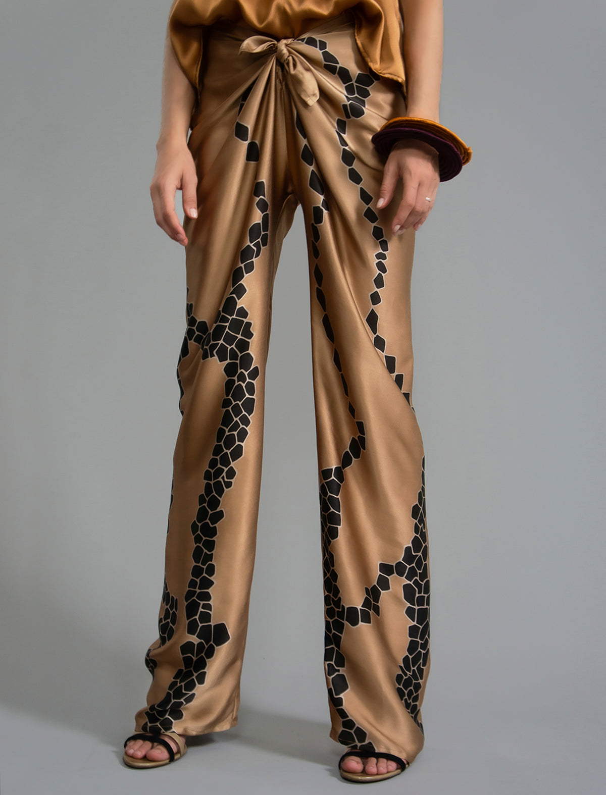 Front detail view. Long silk pants in tobacco colour with contrasting blue graphic Macrosnake batik motif, featuring a knot tie at the front and a slick relaxed silhouette. 