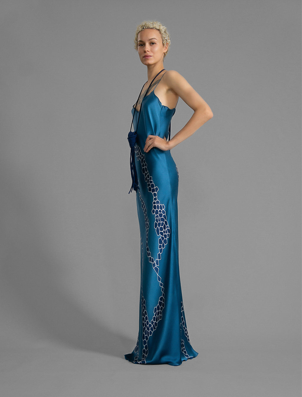 Side view. Elegant long body contouring silk dress in saxony blue colour with contrasting blue graphic Macrosnake batik motif, featuring spaghetti straps, V-neckline and a fluid contemporary silhouette.