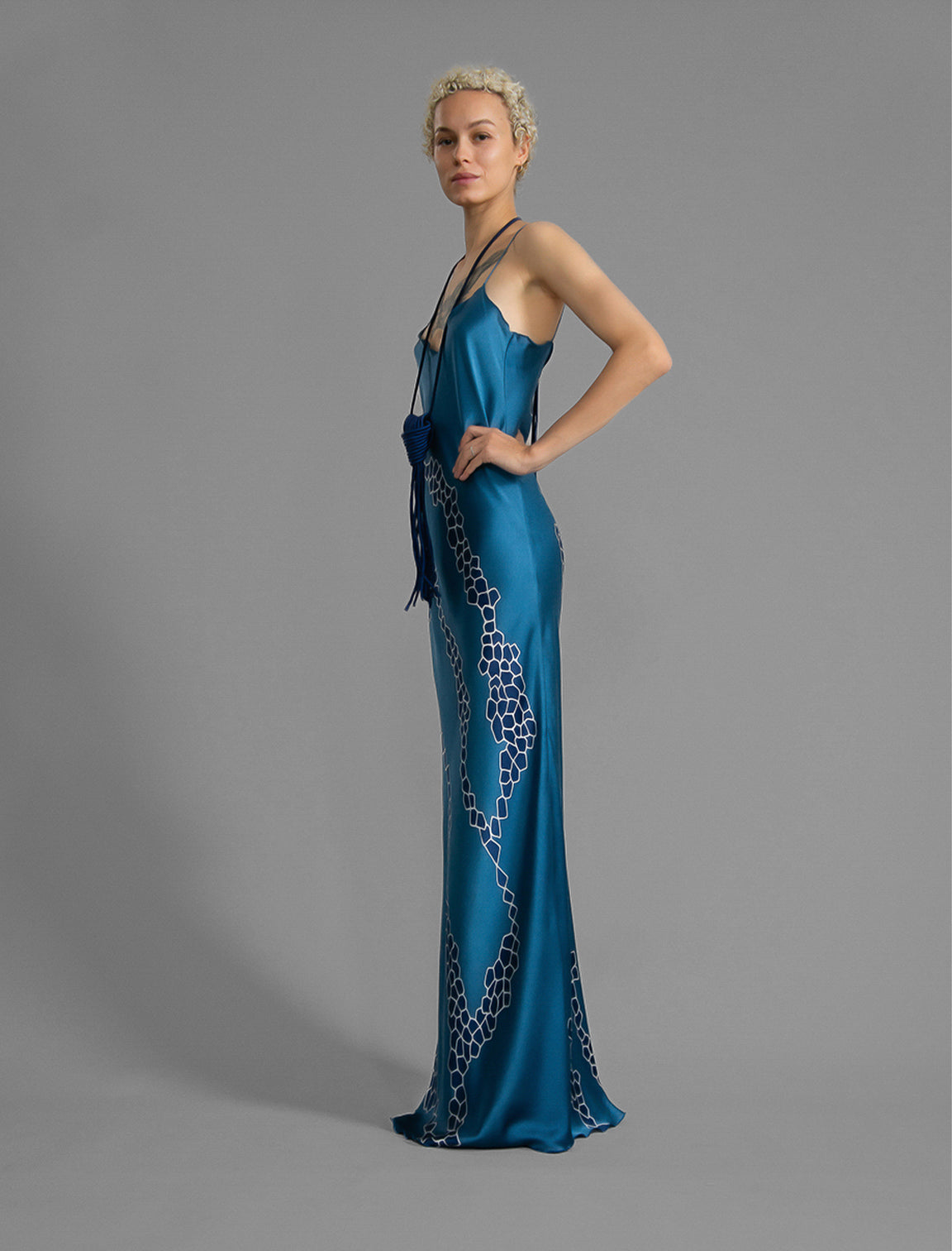 Side view. Elegant long body contouring silk dress in saxony blue colour with contrasting blue graphic Macrosnake batik motif, featuring spaghetti straps, V-neckline and a fluid contemporary silhouette.