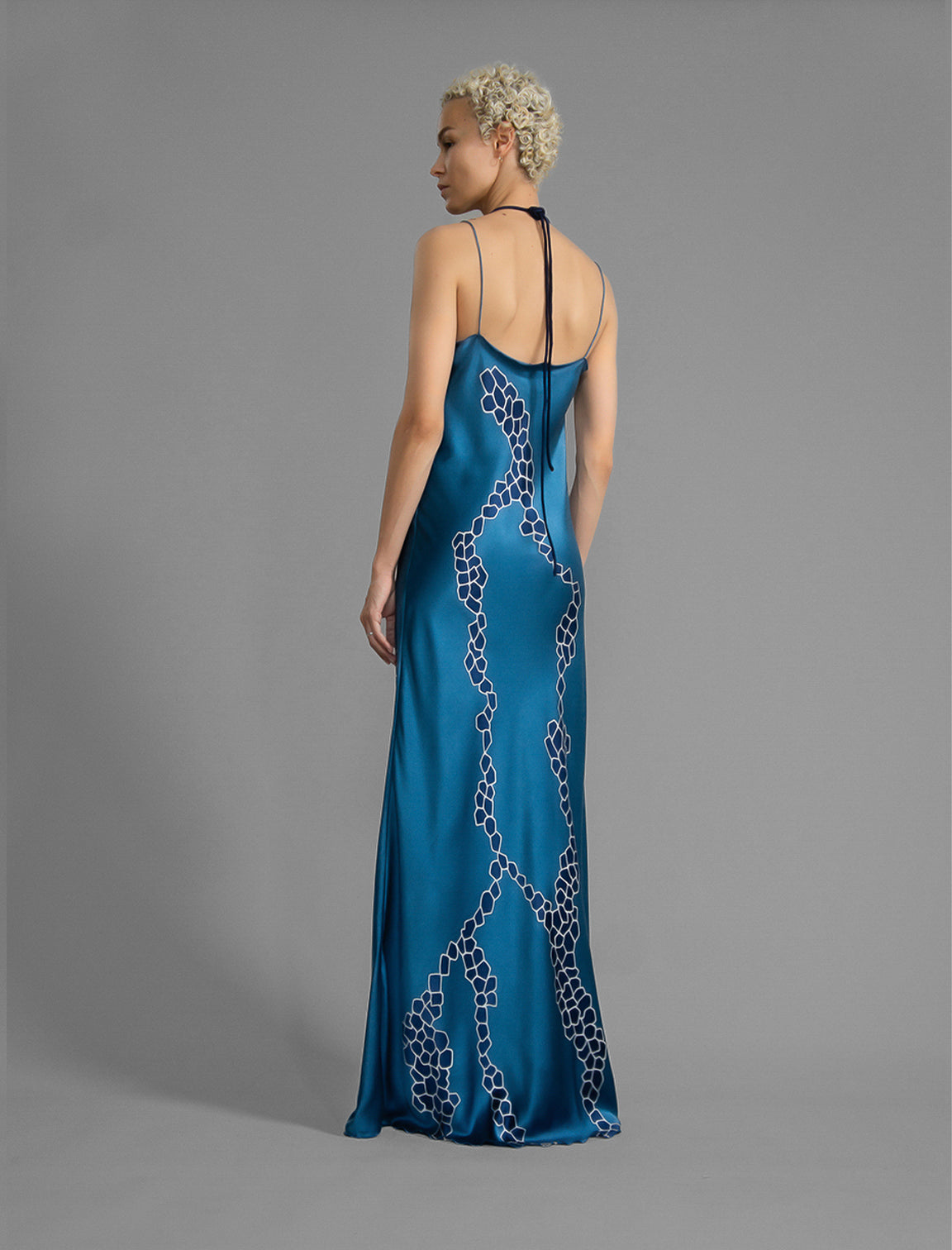 Back view. Elegant long body contouring silk dress in saxony blue colour with contrasting blue graphic Macrosnake batik motif, featuring spaghetti straps, V-neckline and a fluid contemporary silhouette.