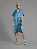 Front view. Mid length shirt dress in dream blue colour with contrasting blue graphic Macrosnake batik motif, featuring short sleeves with deep hem, curved hemline, simple collar with narrow V-neckline and relaxed contemporary silhouette.