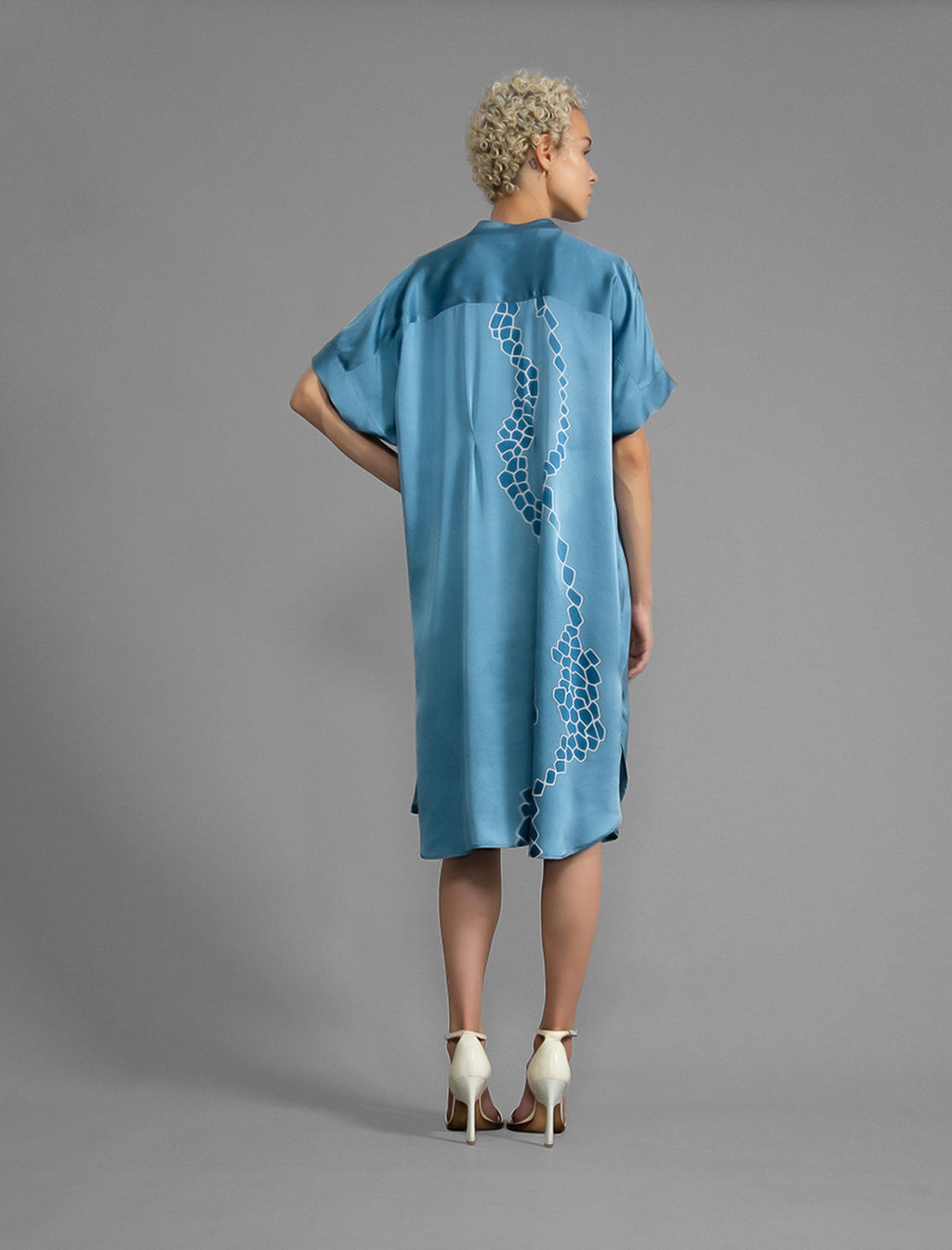 Back view. Mid length shirt dress in dream blue colour with contrasting blue graphic Macrosnake batik motif, featuring short sleeves with deep hem, curved hemline, simple collar with narrow V-neckline and relaxed contemporary silhouette.