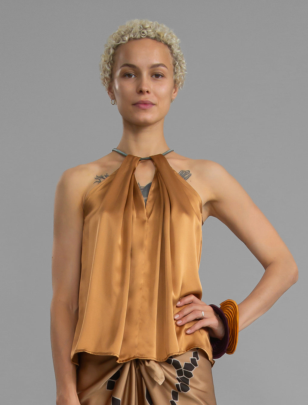Close front view. Silk satin halter top in warm camel colour with elegant draping down the front, a high neckline anchored by a thin, grey silk cord that threads through the fabric, creating a V-shaped opening above the chest. Also draped at the back, the cord ties into a bow over a V-shaped opening. The hemline flows loosely over the hips.