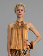 Close front view. Silk satin halter top in warm camel colour with elegant draping down the front, a high neckline anchored by a thin, grey silk cord that threads through the fabric, creating a V-shaped opening above the chest. Also draped at the back, the cord ties into a bow over a V-shaped opening. The hemline flows loosely over the hips.