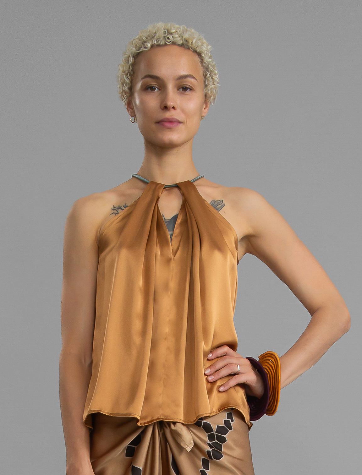 Close front view. Silk satin halter top in warm camel colour with elegant draping down the front, a high neckline anchored by a thin, grey silk cord that threads through the fabric, creating a V-shaped opening above the chest. Also draped at the back, the cord ties into a bow over a V-shaped opening. The hemline flows loosely over the hips.