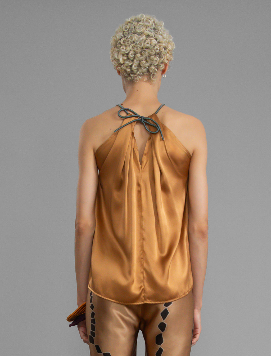 Close back view. Silk satin halter top in warm camel colour with elegant draping down the front, a high neckline anchored by a thin, grey silk cord that threads through the fabric, creating a V-shaped opening above the chest. Also draped at the back, the cord ties into a bow over a V-shaped opening. The hemline flows loosely over the hips.