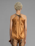 Close back view. Silk satin halter top in warm camel colour with elegant draping down the front, a high neckline anchored by a thin, grey silk cord that threads through the fabric, creating a V-shaped opening above the chest. Also draped at the back, the cord ties into a bow over a V-shaped opening. The hemline flows loosely over the hips.