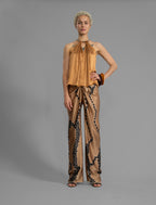 Front view. Silk satin halter top in warm camel colour with elegant draping down the front, a high neckline anchored by a thin, grey silk cord that threads through the fabric, creating a V-shaped opening above the chest. Also draped at the back, the cord ties into a bow over a V-shaped opening. The hemline flows loosely over the hips.