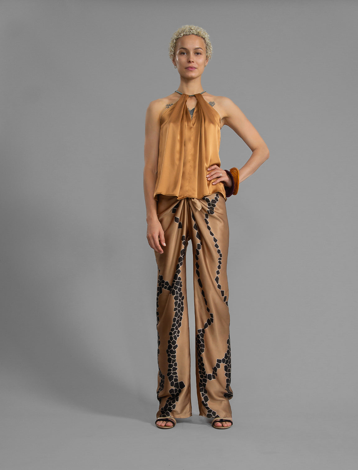 Front view. Silk satin halter top in warm camel colour with elegant draping down the front, a high neckline anchored by a thin, grey silk cord that threads through the fabric, creating a V-shaped opening above the chest. Also draped at the back, the cord ties into a bow over a V-shaped opening. The hemline flows loosely over the hips.