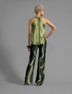 Back view. Silk satin halter top in fern green colour with elegant draping down the front, a high neckline anchored by a thin, grey silk cord that threads through the fabric, creating a V-shaped opening above the chest. Also draped at the back, the cord ties into a bow over a V-shaped opening. The hemline flows loosely over the hips.