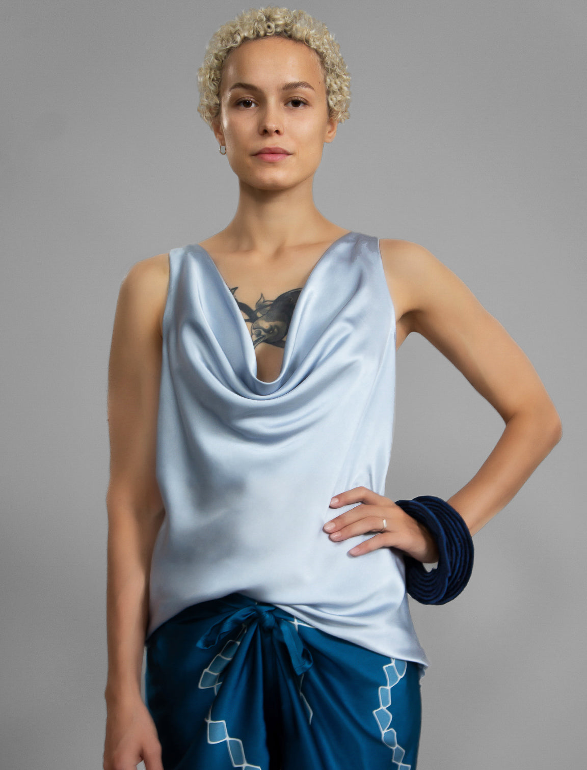 Close view. Sleeveless silk top in pale ice blue with glossy satin finish, featuring a dramatic, deeply draped cowl neckline that creates elegant, fluid folds across the chest. It has wide, comfortable shoulder straps and a relaxed silhouette that flows gently down to a hemline over the hips.