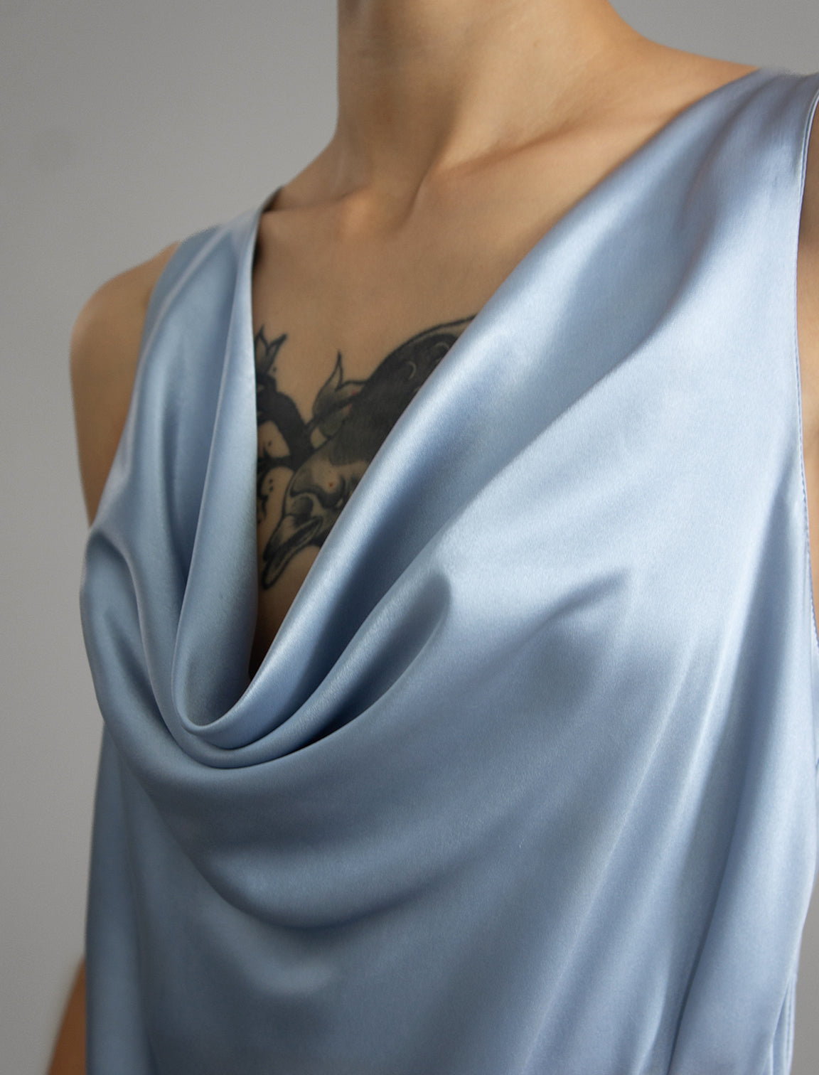 Neckline detail: Sleeveless silk top in pale ice blue with glossy satin finish, featuring a dramatic, deeply draped cowl neckline that creates elegant, fluid folds across the chest. It has wide, comfortable shoulder straps and a relaxed silhouette that flows gently down to a hemline over the hips.