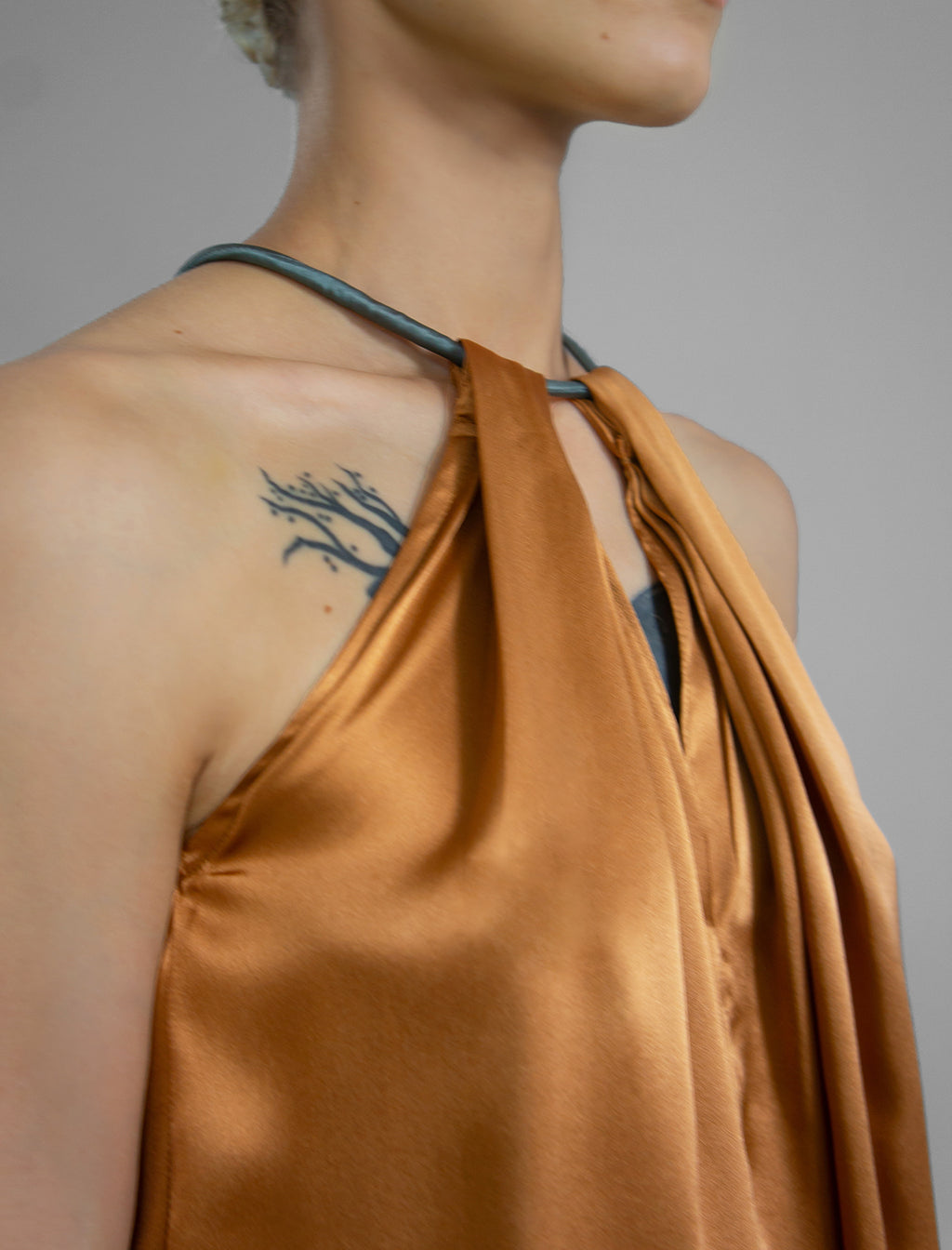 Front detail: Silk satin halter top in warm camel colour with elegant draping down the front, a high neckline anchored by a thin, grey silk cord that threads through the fabric, creating a V-shaped opening above the chest. Also draped at the back, the cord ties into a bow over a V-shaped opening. The hemline flows loosely over the hips.