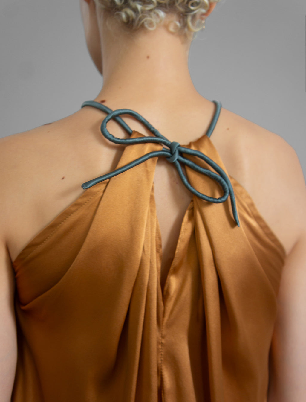 Back detail: Silk satin halter top in warm camel colour with elegant draping down the front, a high neckline anchored by a thin, grey silk cord that threads through the fabric, creating a V-shaped opening above the chest. Also draped at the back, the cord ties into a bow over a V-shaped opening. The hemline flows loosely over the hips.