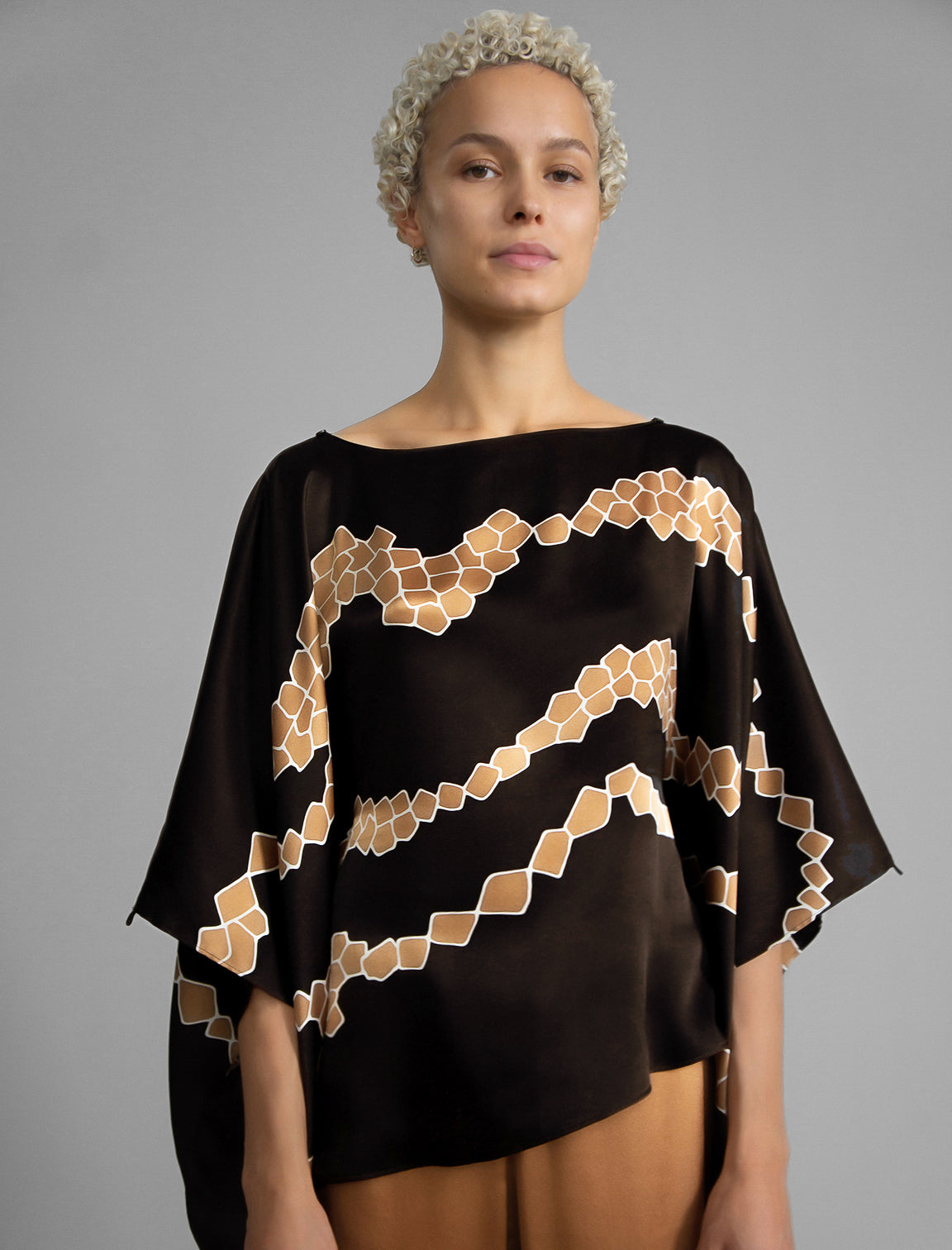 Front view. Flowing coffee colour silk top with graphic Macrosnake batik motif in warm beige tones, featuring fluid draped sleeves, asymmetrical hemline, and versatile contemporary silhouette designed to be worn three ways.