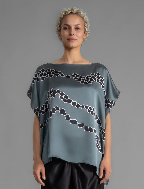 Front view. Flowing steel grey colour silk top with graphic Macrosnake batik motif in contrasting black, featuring fluid draped sleeves, asymmetrical hemline, and versatile contemporary silhouette designed to be worn three ways.