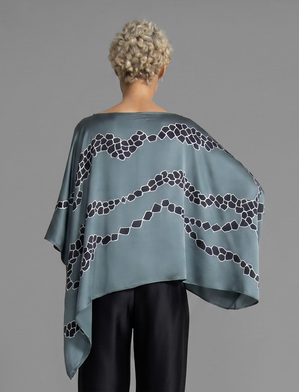 Back view. Flowing steel grey colour silk top with graphic Macrosnake batik motif in contrasting black, featuring fluid draped sleeves, asymmetrical hemline, and versatile contemporary silhouette designed to be worn three ways.