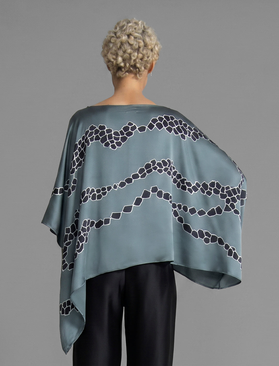 Back view. Flowing steel grey colour silk top with graphic Macrosnake batik motif in contrasting black, featuring fluid draped sleeves, asymmetrical hemline, and versatile contemporary silhouette designed to be worn three ways.