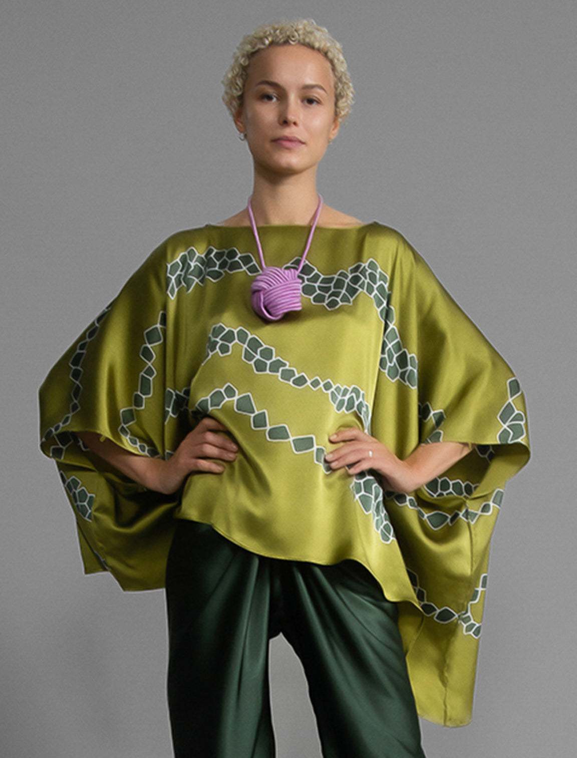 Front view. Flowing moss green colour silk top with graphic Macrosnake batik motif in contrasting green tone, featuring fluid draped sleeves, asymmetrical hemline, and versatile contemporary silhouette designed to be worn three ways.